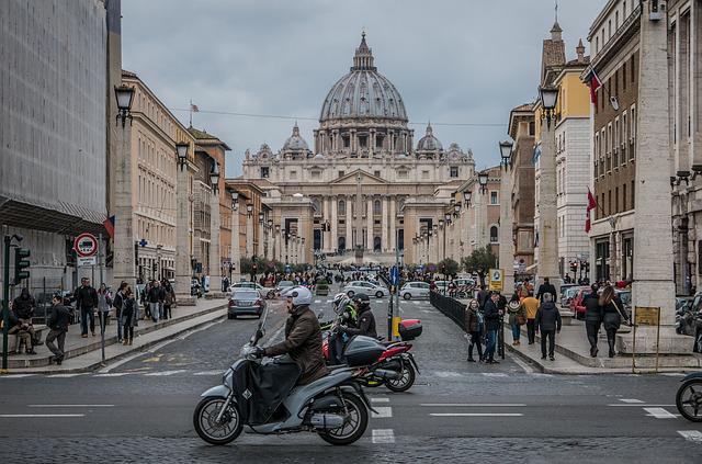 Bild på Peterskyrkan i Vatikanstaten i Rom med surrande trafik i förgrunden bestående av mopedister, bilar och gångtrafikanter.